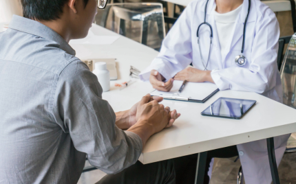 Healthcare professional consulting with a patient during an in-person appointment, discussing medical information across a desk with a tablet and clipboard visible.