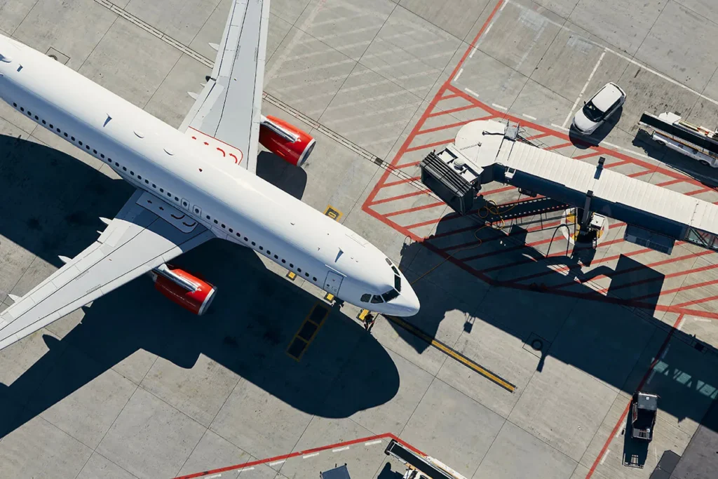 Aerial view of a passenger airplane parked at an airport gate with a jet bridge and ground service vehicles.