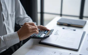 A person using a calculator while reviewing printed financial documents at a desk. A clipboard with charts and tables sits beside a closed laptop, suggesting budgeting, accounting, or financial analysis in a modern office setting.